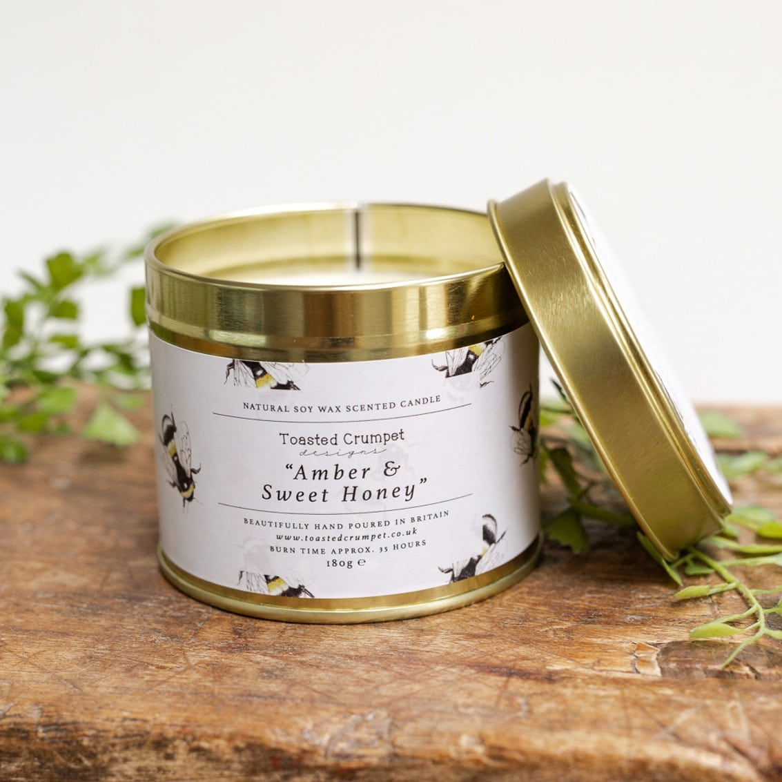 Candle tin labeled 'Amber & Sweet Honey' on a wooden surface with greenery in the background
