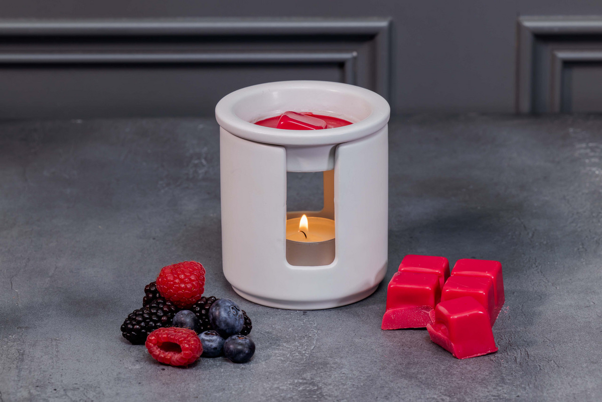 White wax warmer with a lit candle surrounded by berries and red wax cubes on a gray surface.